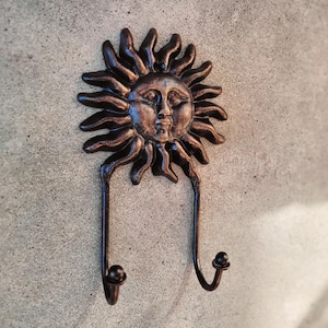 Puede incluir: Un gancho de pared de hierro fundido con forma de sol con cara. El gancho tiene dos puntas para colgar objetos. El sol es de color marrón oscuro.