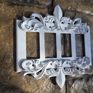 Könnte beinhalten: Weiße dekorative Lichtschalterabdeckung mit aufwendigem Fleur-de-Lis- und Rankenwerk-Design. Die Abdeckung hat vier rechteckige Öffnungen und ist auf einer strukturierten, braunen Oberfläche montiert. Das Design hat schwarze Akzente.