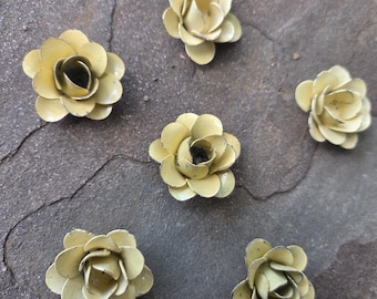 Puede incluir: Seis pequeños adornos florales de metal amarillo con un acabado envejecido están dispersos sobre una superficie oscura y texturizada. Las flores tienen múltiples pétalos y un centro oscuro, lo que sugiere una estética vintage o artesanal.