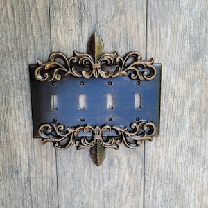 Może przedstawiać: Dekoracyjny włącznik światła z czarnym tłem i ozdobną, złoconą metalową ramką. Ramka zdobiona jest motywem lilii i misternymi zdobieniami.