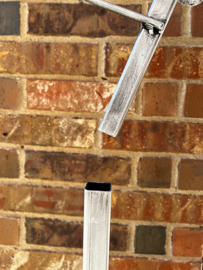 May include: Close-up of a silver metal stand with a square base and a rectangular top. The stand is made of metal tubing and has a brushed finish.
