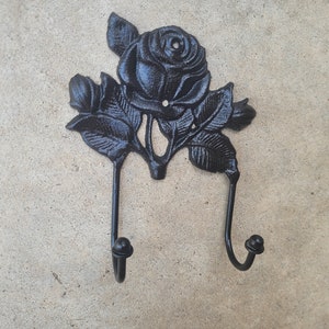 May include: Black cast iron wall hook featuring a rose design with two hooks. The decorative hook is in the shape of a rose with leaves and two curved hooks below. The hook is approximately 6 inches wide.