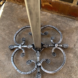 May include: A silver metal stand with a decorative base featuring a swirling design with leaf accents. The stand is designed to hold a variety of items, such as a plant or a decorative object.