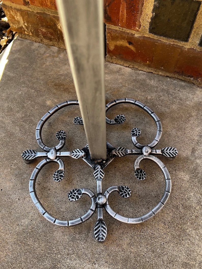 May include: A silver metal stand with a decorative base featuring a swirling design with leaf accents. The stand is designed to hold a variety of items, such as a plant or a decorative object.