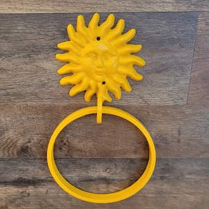 May include: Yellow metal sun-shaped towel ring with a face on the sun. The ring is attached to a wooden wall.