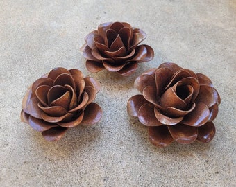 Puede incluir: Tres esculturas de flores decorativas individuales hechas de metal marrón. Cada pieza presenta pétalos curvos y superpuestos dispuestos en un patrón circular de rosa en flor con un acabado texturizado y un color marrón intenso y profundo.