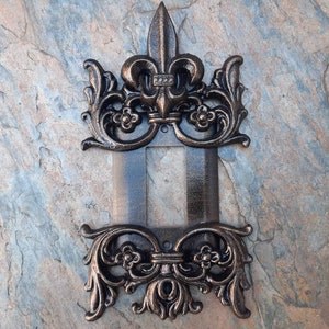 Puede incluir: Una cubierta de interruptor de luz decorativa de hierro fundido con un diseño de flor de lis en acabado bronce.