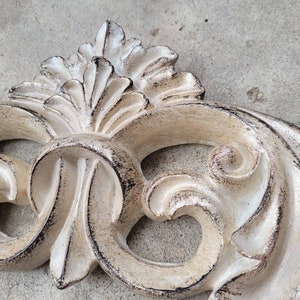 May include: A white, ornate, decorative wall decor piece with a distressed finish. The piece is made of plaster or resin and features a swirling, floral design.