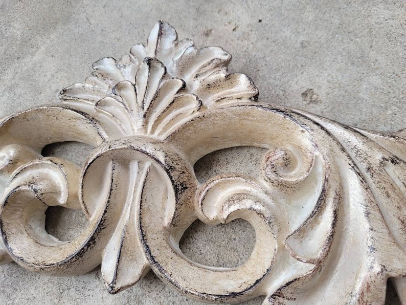 May include: A white, ornate, decorative wall decor piece with a distressed finish. The piece is made of plaster or resin and features a swirling, floral design.