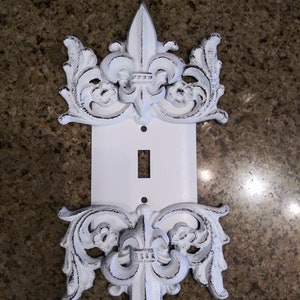May include: White painted metal light switch cover with a decorative fleur-de-lis design on the top and bottom.
