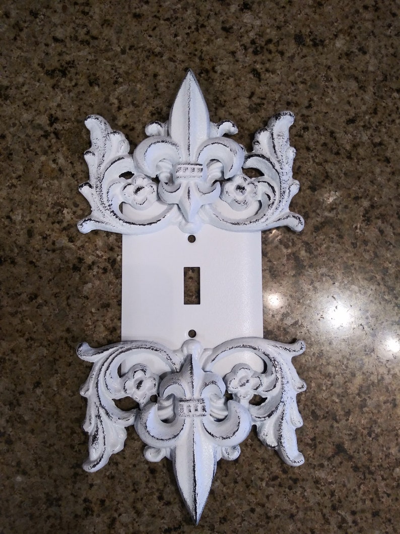 May include: White painted metal light switch cover with a decorative fleur-de-lis design on the top and bottom.