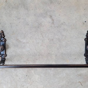 May include: Black metal towel rack with ornate scrollwork and knob accents. The rack is mounted on a wall.
