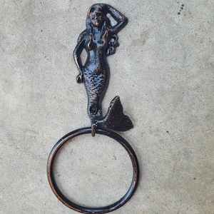 May include: Black metal towel ring with a mermaid design. The mermaid is holding her hand to her head and has a long flowing tail.