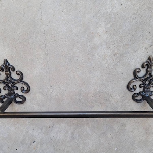 Puede incluir: Toallero ornamental de bronce oscuro con detalles decorativos de flor de lis. La barra de la toalla es una varilla recta y horizontal con extremos redondeados. Montado en una pared de hormigón gris claro.