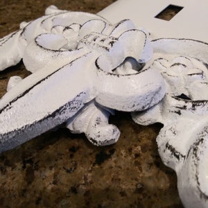 May include: White decorative switch plate covers with an ornate design. The covers have a distressed look with black paint showing through the white paint.