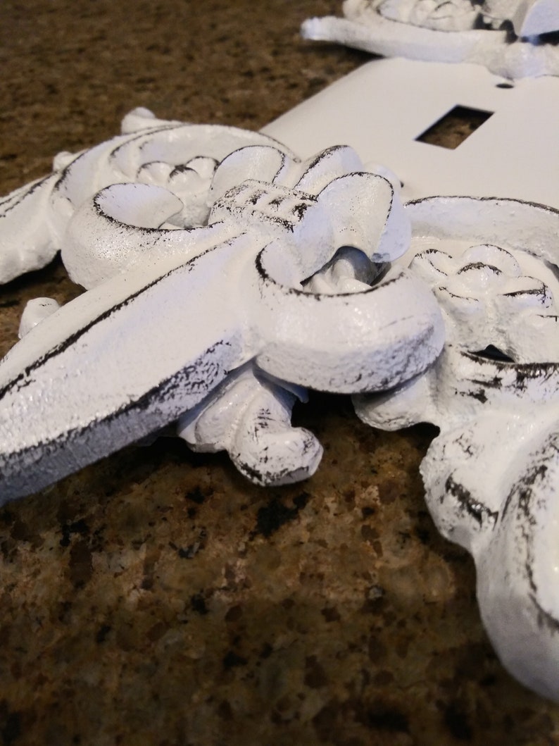 May include: White decorative switch plate covers with an ornate design. The covers have a distressed look with black paint showing through the white paint.