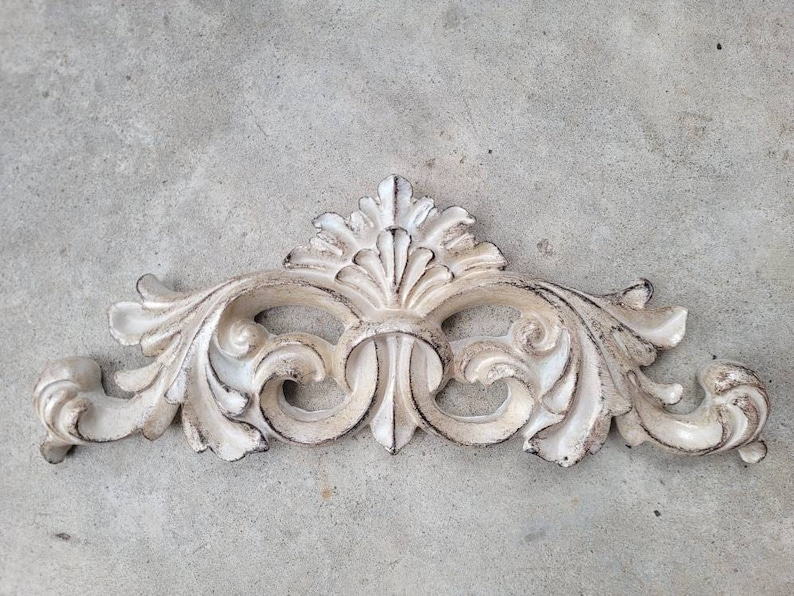 May include: A white, ornate, decorative wall accent with a scroll-like design. The accent is made of plaster or a similar material and has a distressed, aged look.