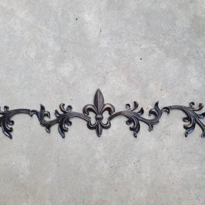 Op de afbeelding: Een donkerbronzen metalen decoratief krulwerk met een leliecentrum. Het symmetrische ontwerp heeft krullende bladeren en rondingen, geschikt voor meubels of wanddecoratie. Het stuk is geplaatst tegen een neutrale grijze achtergrond.