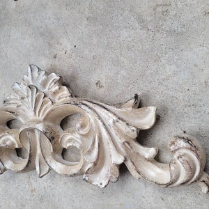 May include: A white, ornate, decorative wall decor piece with a distressed finish. The piece is made of wood and has a floral design.