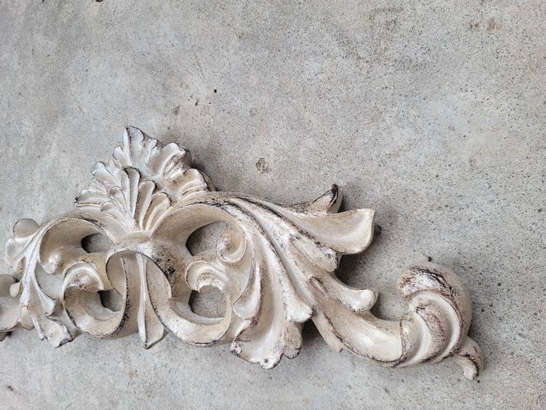 May include: A white, ornate, decorative wall decor piece with a distressed finish. The piece is made of wood and has a floral design.