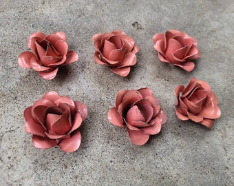 Puede incluir: Seis flores de rosas de metal hechas a mano en un color rosa polvoriento apagado. Las flores están dispuestas sobre una superficie de hormigón gris. Cada rosa tiene múltiples pétalos y el metal tiene un acabado ligeramente envejecido.