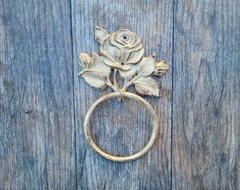 May include: A gold-toned metal towel ring with a decorative rose design. The ring is mounted on a wooden surface.
