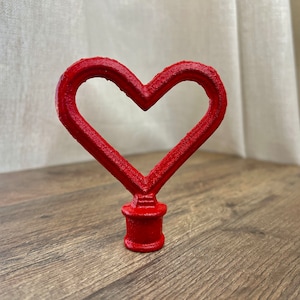 May include: A red metal heart-shaped finial with a smooth, glossy finish. The finial is designed to be used as a decorative topper for furniture or other objects.