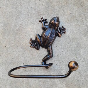 May include: A black metal frog figurine with a bronze finish, holding a black metal toilet paper holder with a round knob.