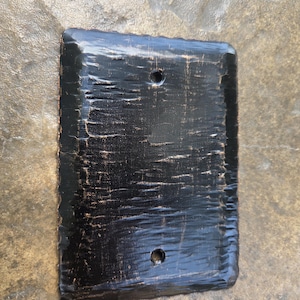 May include: A rectangular wall plate cover featuring a textured, hammered black finish with subtle copper-colored highlights along the edges and surface. The plate includes two circular screw holes positioned vertically at the top and bottom center.