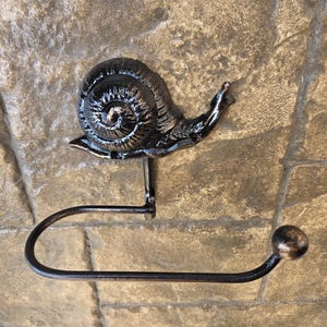 May include: A black metal toilet roll holder with a snail design. The snail has a textured shell, and the holder includes a curved bar for the roll. The holder is mounted on a stone wall. The overall design is rustic.
