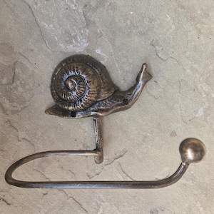 May include: Bronze-colored metal snail toilet paper holder. The snail has a detailed shell and a curved body. The holder has a curved bar for the toilet paper roll and a ball at the end. The holder is designed to be mounted on a wall.
