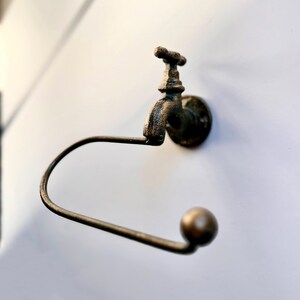 May include: A rustic-style toilet paper holder with a faucet design. The holder is made of metal and has a dark brown finish. The faucet has a round knob and a spout that curves downward.