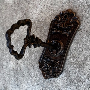 Op de afbeelding: Ouderwetse sleutel en rozet in bronskleurige afwerking. De sleutel heeft een decoratieve handgreep en de rozet heeft gedetailleerde bloemen en krullen. Een vintage-ge&iuml;nspireerd decoratief element.