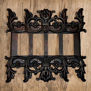 Könnte beinhalten: Eine schwarze Metall-Schalterplatte mit einem verzierten, dekorativen Design. Die Platte hat drei Öffnungen für Schalter.