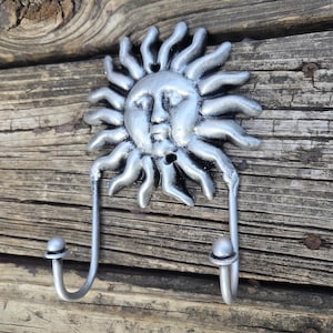 Puede incluir: Gancho de pared de metal plateado con un diseño de cara de sol con rayos radiantes. El gancho tiene dos brazos curvos con extremos redondeados para colgar objetos. Montado sobre una superficie de madera envejecida.