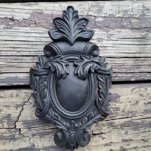 May include: Black ornate decorative wall plaque with a floral design. The plaque has a raised oval center with a smooth surface.