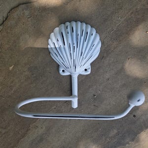 May include: A white metal wall-mounted toilet paper holder featuring a decorative seashell-shaped backplate. The holder has a curved arm with a small ball finial at the end and shows signs of distressed paint revealing dark metal underneath.