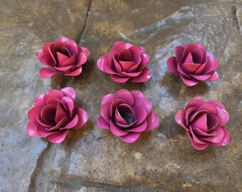 Puede incluir: Seis flores de rosas de metal de color magenta con pétalos detallados están dispuestas sobre una superficie texturizada. Las flores están en varias etapas de floración, mostrando su intrincado diseño y color vibrante.