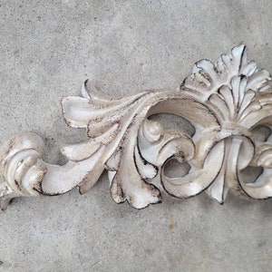 May include: A white, ornate, decorative, plaster, or wood, wall decor piece with a scroll design. The piece has a distressed, aged, or antique finish.