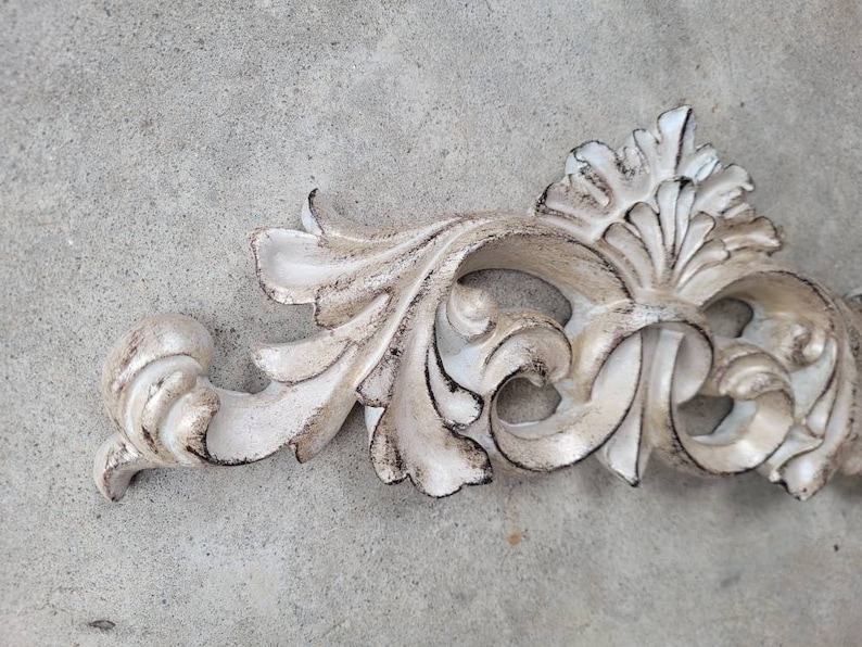 May include: A white, ornate, decorative, plaster, or wood, wall decor piece with a scroll design. The piece has a distressed, aged, or antique finish.