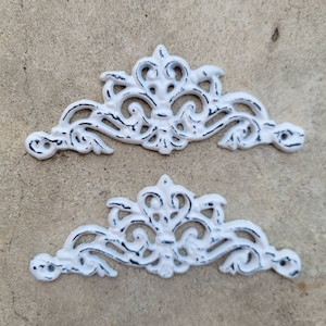 May include: Two white, ornate, metal decorative pieces with a distressed finish. The pieces are shaped like a crown or crest and have a delicate, intricate design.