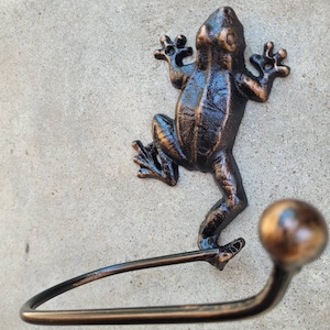 May include: A black metal frog-shaped towel holder with a bronze finish. The frog is holding a metal bar that is curved to hold a towel.