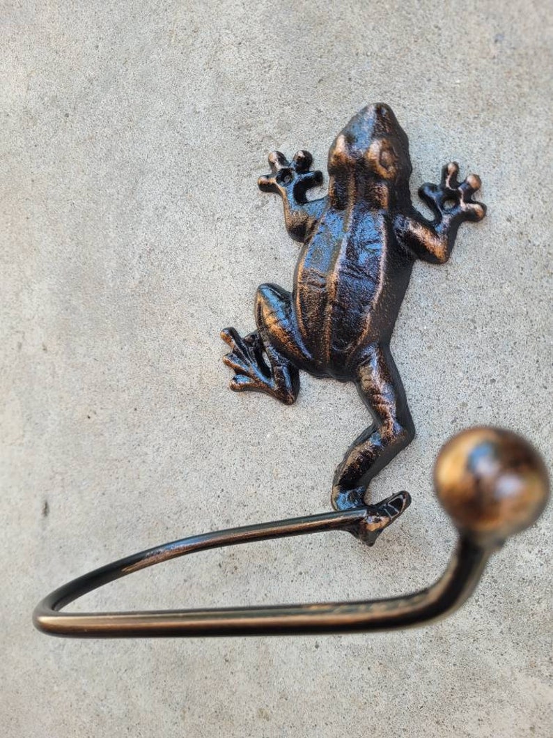 May include: A black metal frog-shaped towel holder with a bronze finish. The frog is holding a metal bar that is curved to hold a towel.