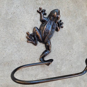 May include: A bronze frog-shaped toilet paper holder with a round knob at the end of the arm. The frog is standing on its hind legs with its front legs raised.