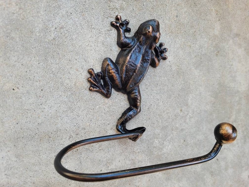 May include: A bronze frog-shaped toilet paper holder with a round knob at the end of the arm. The frog is standing on its hind legs with its front legs raised.