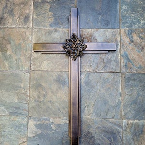 May include: A dark brown wooden cross with a decorative floral design in the centre. The cross is mounted on a wall with a slate tile background.