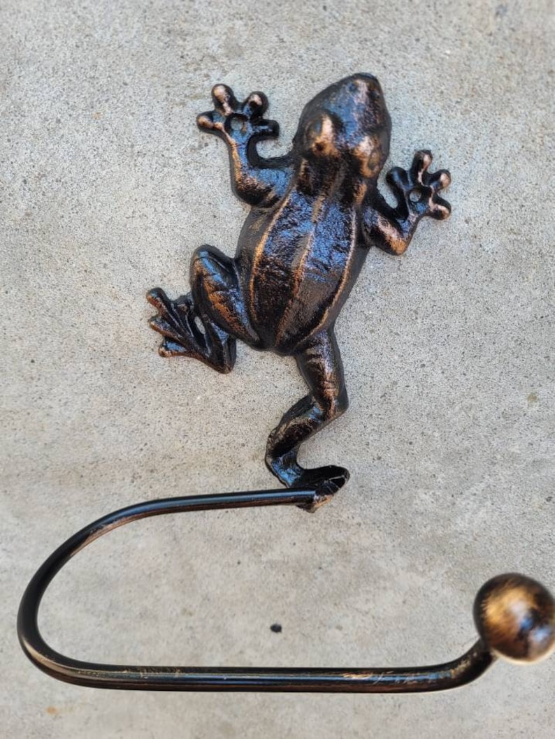 May include: A black metal frog-shaped toilet paper holder with a gold-colored accent on the end of the roll holder.