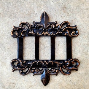 May include: A black and gold metal wall plate with three rectangular openings. The plate is decorated with ornate scrollwork and fleur-de-lis designs.