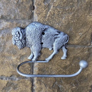 May include: A silver-colored metal bison toilet paper holder. The bison is detailed with textured fur and has a curved arm for holding the toilet paper roll. The holder is mounted on a stone wall.