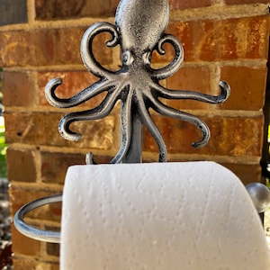 May include: Silver octopus-shaped toilet paper holder with a roll of white toilet paper. The holder is mounted on a brick wall.